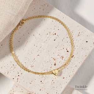 K&ouml;nnte beinhalten: Ein goldenes Fu&szlig;kettchen mit einem kleinen Herz-Anh&auml;nger mit dem Buchstaben L. Das Fu&szlig;kettchen liegt auf einer hellbeigen, strukturierten Oberfl&auml;che. Twinkle Personalisiert.