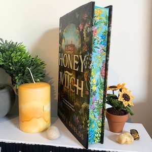 Op de afbeelding: Een hardcover boek getiteld "The Honey Witch" met een bloemenontwerp en een blauw en bloemenpatroon. Een gele kaars, kleine zonnebloemen in potten en decoratieve stenen zijn ook zichtbaar.