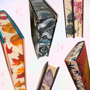 Könnte beinhalten: Eine Sammlung dekorativer Bücher mit bemalten Kanten. Die Bücher zeigen verschiedene Designs, darunter Herbstblätter, Blumenmuster und Himmelsmotive. Die Kanten sind mit leuchtenden Farben bemalt und verleihen jedem Buch eine einzigartige künstlerische Note.