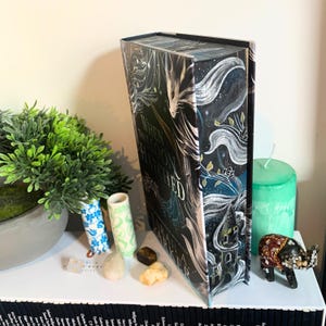 May include: A hardback book titled "When They Moon Hatched" with a dark cover featuring a fantasy illustration of a castle, flowing white shapes, and text. The book is displayed on a shelf with decorative items.