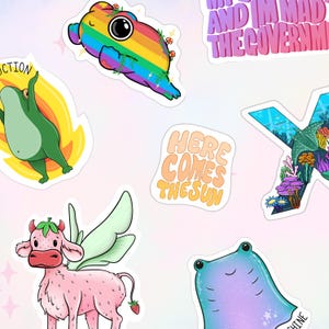 Op de afbeelding: Een verzameling kleurrijke stickers met grillige ontwerpen. Inclusief een regenboogkleurig wezen, een kikker in vlammen, een aardbeienkoe met vleugels en een manta. Tekst bevat "HERE COMES THE SUN" en "AND I'M MAD AT THE GOVERNMENT".