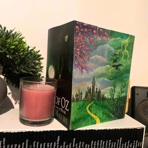 Puede incluir: Una portada de libro pintada a mano para "Wicked: The Untold Story of the Witches of Oz" de Gregory Maguire. La obra de arte representa escenas del libro, incluyendo un camino de ladrillos amarillos, un castillo y un cielo verde con relámpagos. Una vela rosa está frente al libro.