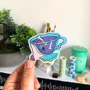 Op de afbeelding: Een holografische sticker met een theekopje met paarse vloeistof en botten erin. De theekop heeft een oor en een schotel. Een theezakje met een schedel is aan de kop bevestigd. De sticker heeft een glanzende, iriserende afwerking.