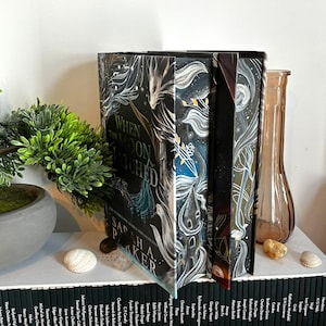 May include: A hardcover book titled "When the Moon Hatched" by Sarah A. Parker. The book has a dark cover with a celestial design in white and blue. The spine is black with white text. The book is displayed with other books and decorative items.