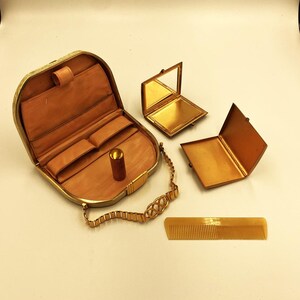 May include: An antique gold-toned vanity set, including a compact mirror, a small case, a lipstick, a comb, and a small purse with a chain. The set is displayed on a white surface. The purse is open, revealing a tan interior.
