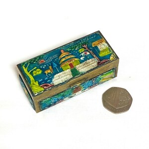 May include: A rectangular, vintage trinket box with a detailed, colourful enamel design. The box features a turquoise background with a landscape scene, including a building, trees, and a deer. The box is made of metal with a gold finish.