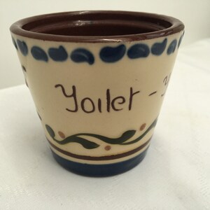 May include: A white ceramic pot with blue and brown accents. The pot has a brown rim and the words "Yoilet -" written on the front.