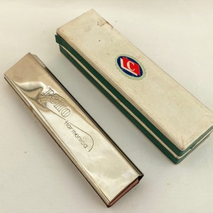 May include: A silver harmonica in a white box with green trim. The harmonica has the word "ECHO Harmonica" engraved on the top. The box has a red and blue logo with the letter "C" in the center.