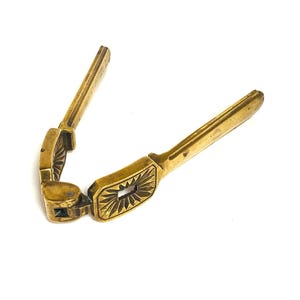 May include: A vintage brass nutcracker, open to reveal its design. The tool has a hinged mechanism and decorative detailing. The handles and the central cracking section are visible, highlighting the tool's function.