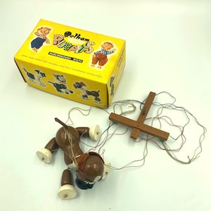 May include: A brown wooden dog marionette with white paws and a wooden control crossbar with strings, accompanied by a yellow cardboard box labelled Pelham Puppets Marlborough Wilts with illustrations of various puppet characters on the exterior.