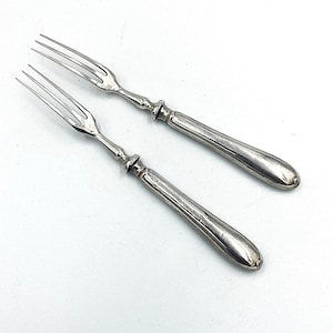 May include: Two silver-toned metal forks with three slender tines and smooth, rounded handles. Each fork features a distinct bolster where the handle meets the neck, showcasing a polished, reflective surface with subtle signs of age and wear.