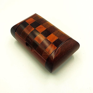 May include: A rectangular wooden box with a checkerboard pattern on the lid. The box is made of various shades of brown wood, with darker wood for the edges and base. It has a curved top and a small hole on the side.