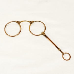 May include: Antique gold-toned pince-nez eyeglasses with round lenses and a handle. The glasses feature a bridge connecting the two lenses and a long, slender handle with a ring at the end. The design is simple and elegant.