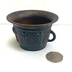 May include: A small, dark brown bronze pot with a wide rim and two handles. The pot has a decorative design of four stylised clover leaves.