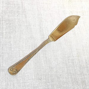 May include: A vintage butter knife with a gold-toned blade and handle. The blade has a curved edge, and the handle features an ornate, detailed design. The knife is approximately 18 cm long.