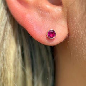 May include: A close-up of an ear with a small stud earring. The earring has a bright pink gemstone set in a gold-coloured bezel. The skin around the ear is a light pink colour, and blonde hair is visible in the background.