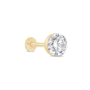 May include: A gold flat back labret stud featuring a round, clear, faceted gemstone set in a gold bezel. The jewelry piece consists of a thin post, a circular flat base, and a prominent, sparkling stone at the front, showcasing a polished finish.