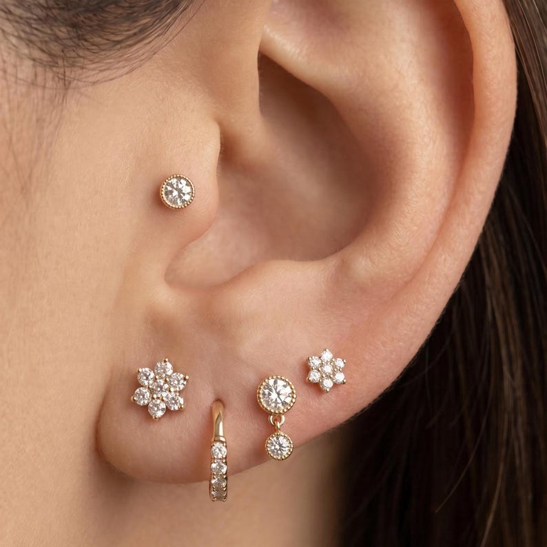 May include: A close-up of an ear adorned with multiple gold earrings. The earrings include a small stud, a flower-shaped stud, a small hoop, and a dangling earring with a circular design. All earrings feature clear, sparkling stones.