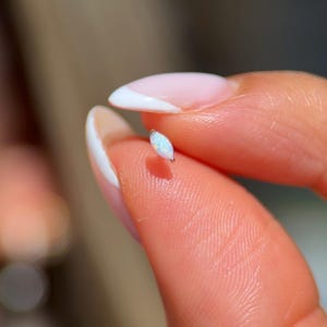 May include: A small, marquise-shaped opal earring with a silver setting. The opal has a pearlescent, iridescent quality, displaying flashes of blue and white. The earring is held between two fingers, showcasing its delicate size.