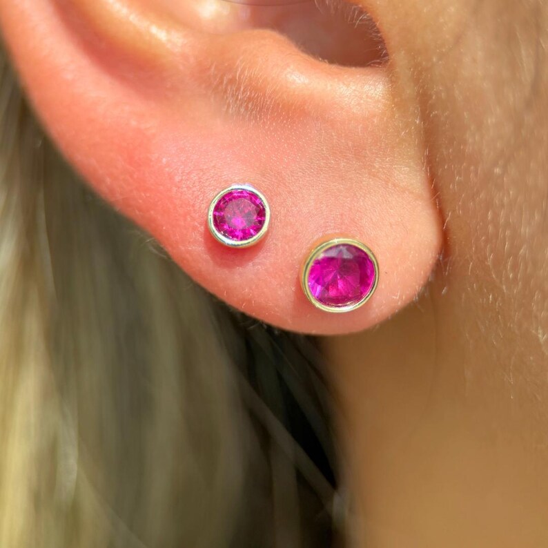 May include: Gold stud earrings featuring bright pink gemstones. The earrings have a round shape with a gold bezel setting. One earring is slightly larger than the other. The earrings are shown on an ear.