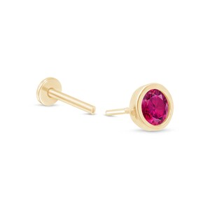 May include: Two gold-coloured labret studs. One stud features a round, bezel-set, ruby-coloured gemstone. The other stud has a flat, circular backing and a straight post. The jewellery is set against a white background.