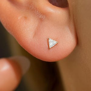 Puede incluir: Primer plano de un pendiente de oro con una gema transparente de forma triangular. El pendiente está engastado en un bisel de oro y se lleva en una oreja. El pendiente es pequeño y delicado.