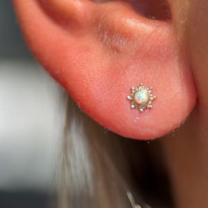 May include: A small, gold-colored stud earring with a central opal stone. The earring has a sunburst design with small, pointed details around the opal. The earring is set in a person's ear.