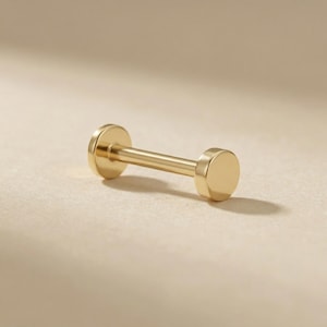 May include: A gold-coloured barbell earring with a flat, circular disc at each end. The earring features a straight, cylindrical bar connecting the two discs. The jewellery is displayed on a neutral, light-coloured surface.