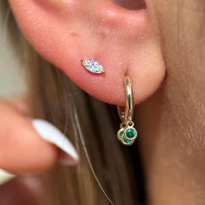 Puede incluir: Primer plano de un pendiente de aro dorado con tres detalles de piedras preciosas verdes y un pendiente de diamante con corte marquesa. Los pendientes están colocados sobre el fondo de una oreja.