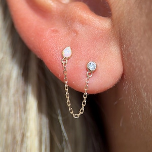 Op de afbeelding: Een paar gouden oorbellen verbonden door een fijne ketting. Eén oorbel heeft een druppelvormige opaal, de andere een kleine, ronde, heldere edelsteen. De oorbellen zijn ontworpen om in aangrenzende oorgaatjes te worden gedragen.