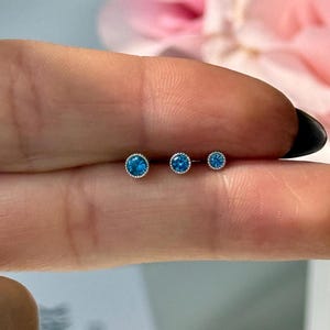 May include: Three silver body jewelry pieces with blue gemstones. The gemstones are round and have a silver bezel setting. The pieces are arranged in order of size, with the smallest on the right and the largest on the left.