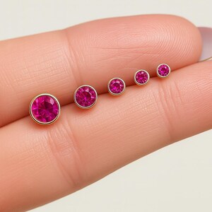 May include: Five gold stud earrings with pink gemstones of varying sizes. The earrings are arranged in a row, with the largest on the left and diminishing in size. The gemstones are round and set in a gold bezel, creating a classic look.