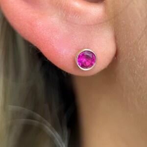 May include: A gold stud earring featuring a bright pink gemstone. The round gemstone is set within a gold bezel, offering a simple, elegant design. The earring is displayed on an ear.