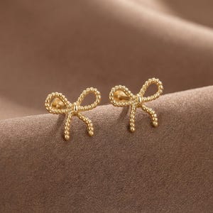May include: A pair of gold-toned stud earrings shaped like bows. Each earring features a textured, rope-like design forming two loops and two dangling ends, with a central knot detail connecting the loops. The earrings have a polished finish.