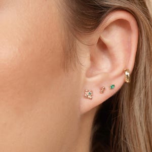 May include: Close-up of a person's ear with multiple earrings. The earrings are gold and feature small green gemstones. One earring is a small, delicate flower design.