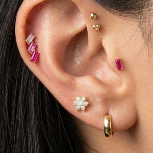 May include: Close-up of an ear adorned with multiple earrings. A gold hoop earring is in the lobe. A small flower-shaped stud with diamonds is above it. A ruby-colored rectangular stud is in the upper ear. Additional gold and ruby earrings are also visible.