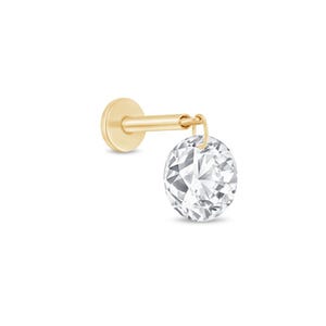 Puede incluir: Un pendiente tipo labret de color dorado con parte posterior plana y un dije colgante de piedra transparente redonda facetada. La pieza consta de un poste de metal liso con una base circular y una pequeña argolla que une la piedra al poste.