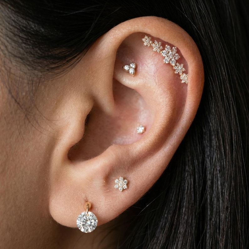 May include: Close-up of an ear adorned with multiple pieces of jewelry. A large, round diamond earring hangs from the lobe. Additional earrings include a cluster of three small diamonds, a single diamond stud, and a floral-shaped earring.