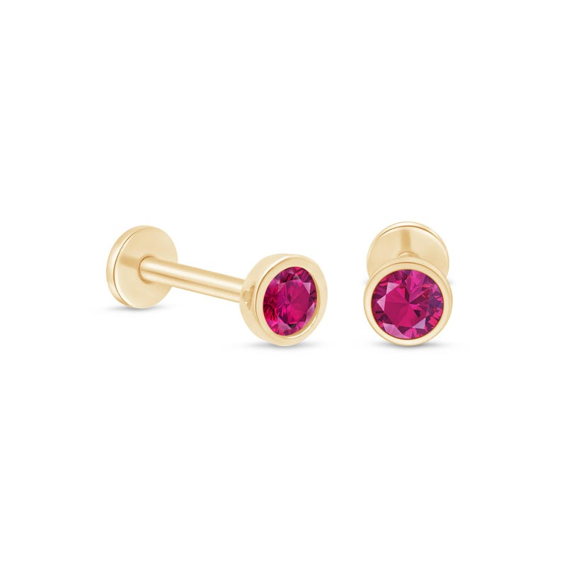 May include: Two gold-coloured, flat-backed earrings with a bright pink gemstone centre. One earring has a post, and the other is a stud. The gemstones are round and set in a bezel setting.