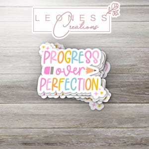 May include: A stack of colorful stickers with the text "PROGRESS over PERFECTION" and a pencil and eraser graphic. The stickers are on a wood background.