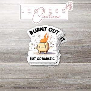 May include: A stack of white stickers with a cartoon marshmallow character on fire, with the words "BURNT OUT" above and "BUT OPTIMISTIC" below. The stickers are on a light wood grain surface.