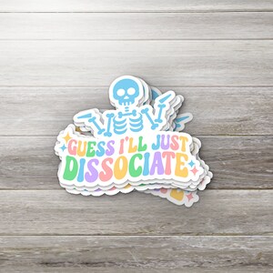 May include: A stack of stickers featuring a light blue skeleton with the text "GUESS I'LL JUST DISSOCIATE" in pastel colors. The stickers have a white border and are on a light wood grain surface.