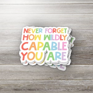 May include: A stack of colorful stickers with the text "NEVER FORGET HOW WILDLY CAPABLE YOU ARE" in a rainbow of colors. The stickers have a white border and are on a wooden surface.
