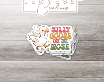 Może przedstawiać: Stos białych naklejek winylowych z kreskówkową gęsią i napisem "SILLY GOOSE ON THE LOOSE" w stylu retro. Naklejki leżą na jasnoszarej drewnianej powierzchni. Marka "LEONESS Creations" znajduje się na górze.