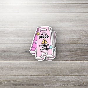 May include: Pink and white caution sign sticker with the text "Not in the mood. Approach with caution" and a yellow exclamation point. The sticker has a cartoon smiley face on each side.