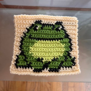May include: A square, crocheted coaster featuring a pixelated frog design. The frog is primarily green with black outlines and a lighter green belly, set against a cream-colored background. The coaster is handmade.