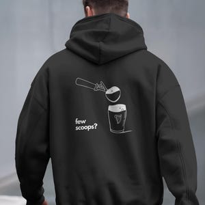 Op de afbeelding: Een zwarte hoodie met een witte afbeelding op de achterkant. Het ontwerp toont een ijsschep boven een pintglas met een harp-logo, vergezeld van de witte tekst "few scoops?" die links van het glas is gedrukt.