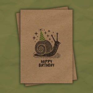 May include: A brown card with a black and white illustration of a snail wearing a green party hat. The snail is surrounded by small white stars. The text "HAPPY BIRTHDAY" is printed below the snail.