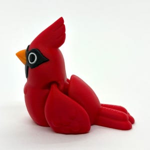 May include: A vibrant red cardinal figurine with a detailed design. The bird has a prominent crest, a black mask around its eye, and an orange beak. The figurine is a decorative item, likely for display.