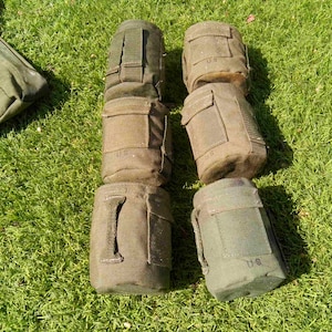 May include: Six olive drab canvas canteen covers arranged in two columns, each featuring a flap closure and the letters US printed on the front. These military-style pouches are constructed from durable fabric with integrated attachment straps.